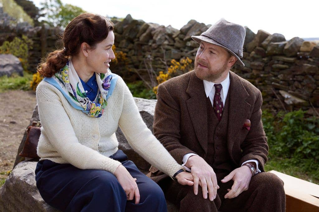Samuel West in a brown suit and hat, sitting on a stone bench with a woman in a white sweater and blue skirt.