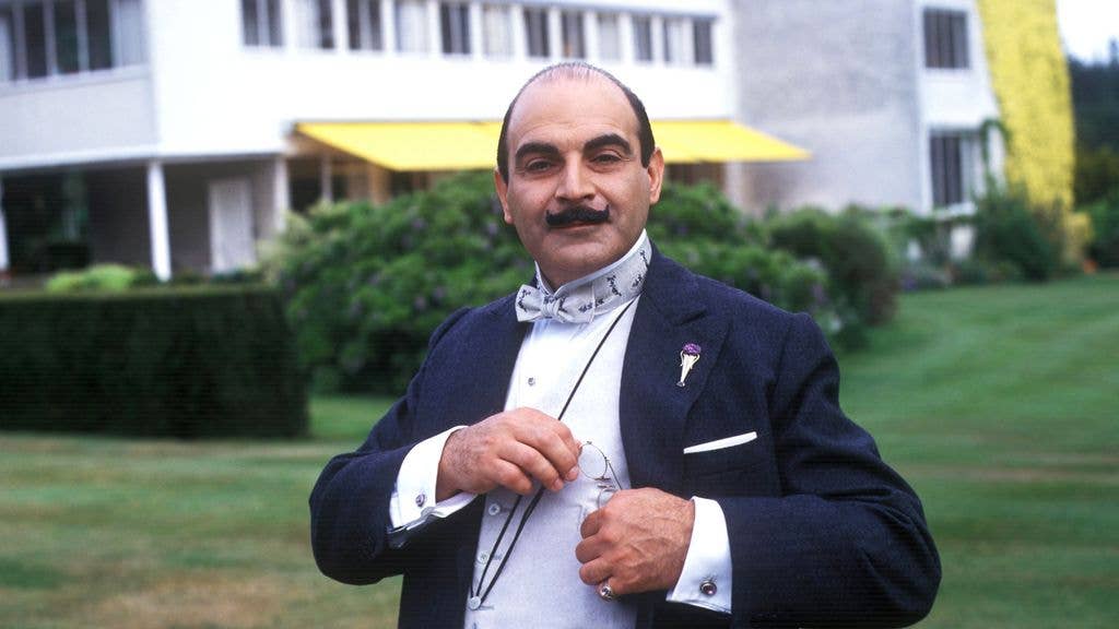 David Suchet in a suit and bow tie, holding glasses, standing in front of a building.