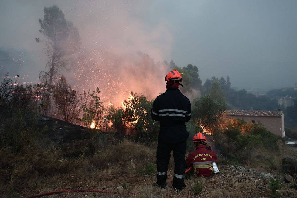 Two firefighters in red helmets and uniforms stand before a wildfire, one seated.