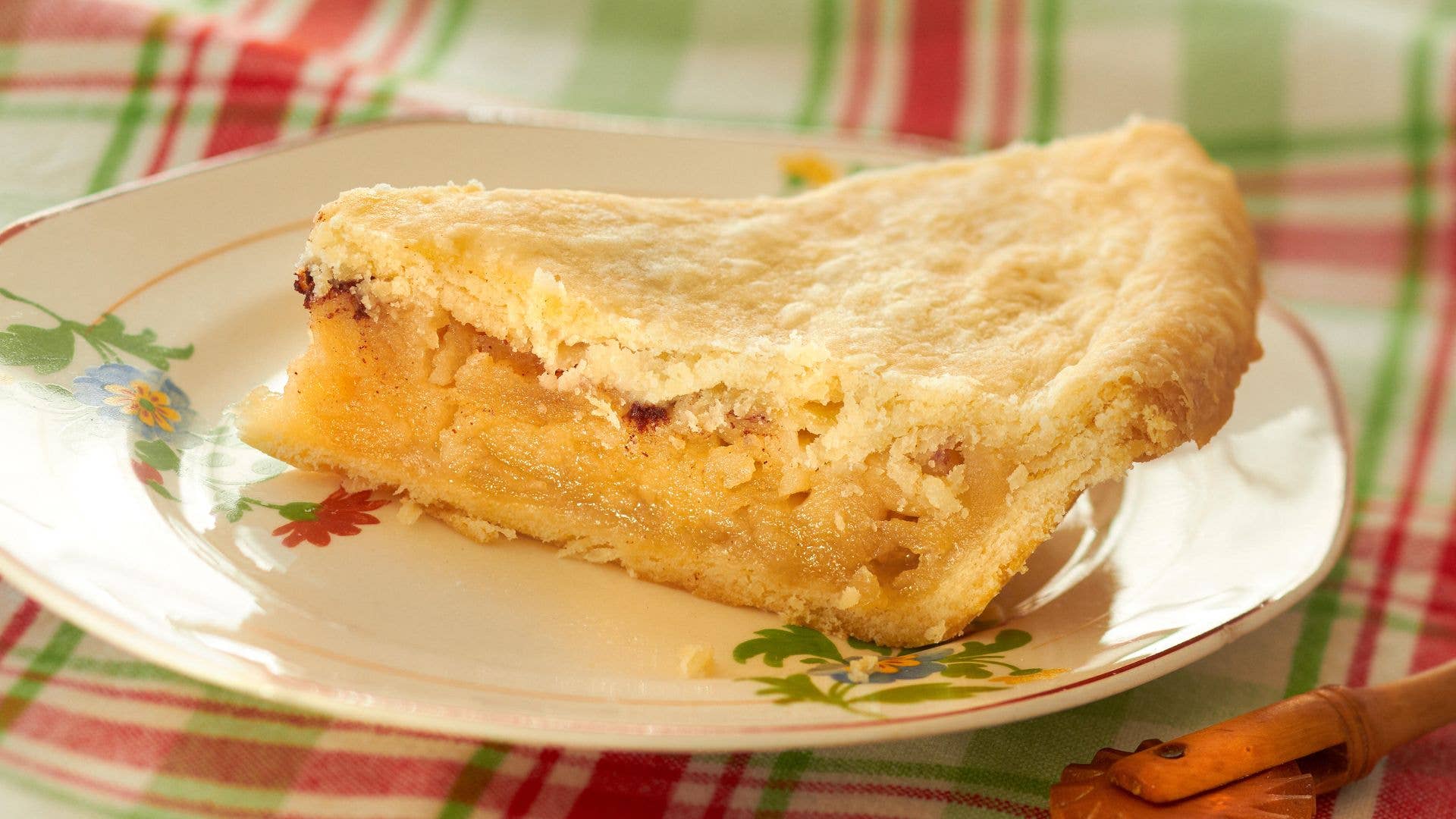 Slice of apple pie on a floral plate, resting on a red and green plaid tablecloth.