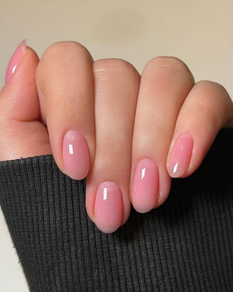 A close-up of a hand with medium-length, oval-shaped nails featuring a "lip gloss" manicure. The polish is a sheer, jelly-like bubblegum pink with a high-gloss, glassy finish that mimics the look of clear lip gloss over natural nails. The hand is posed against a neutral background with a black ribbed sweater sleeve visible.