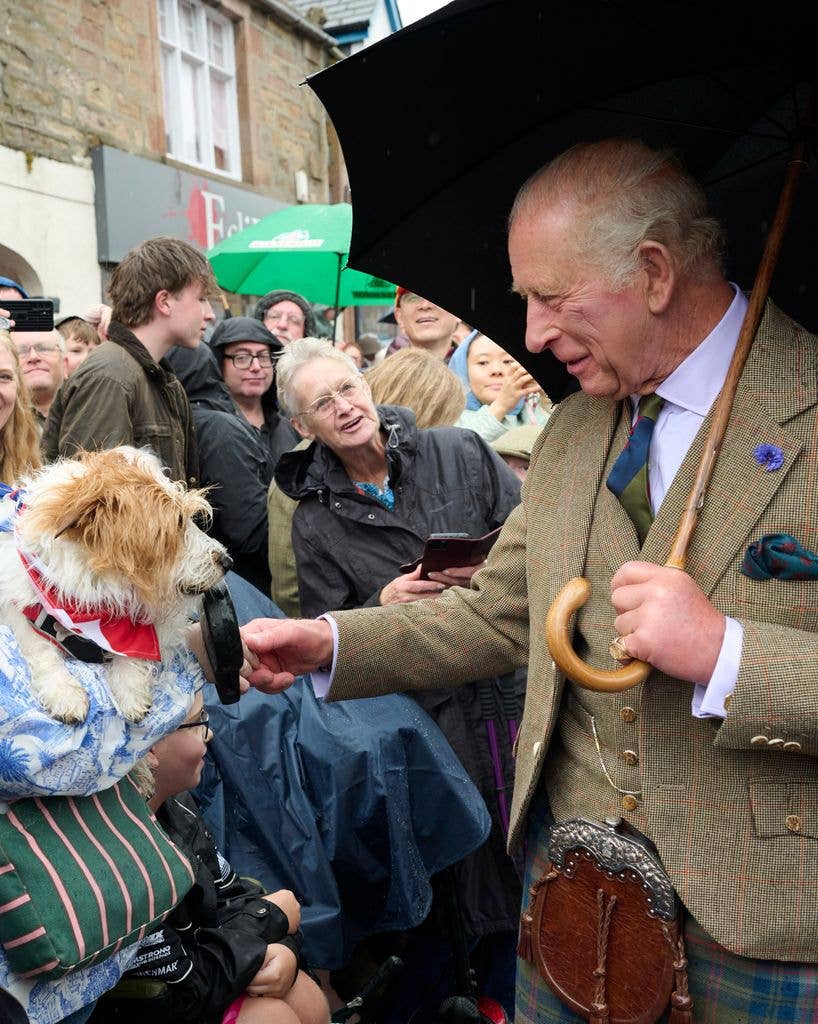 King Charles holding an umbrella, pets a small dog held by a seated person.