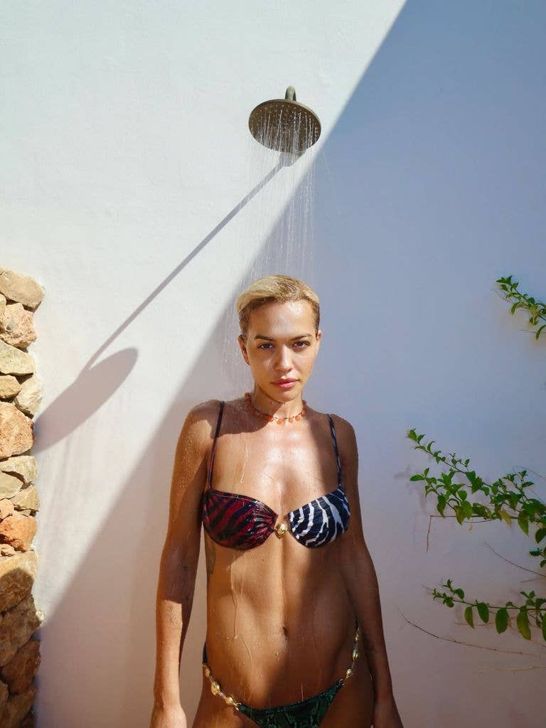 Rita Ora with short blonde hair stands under a shower, wearing a colorful bikini.