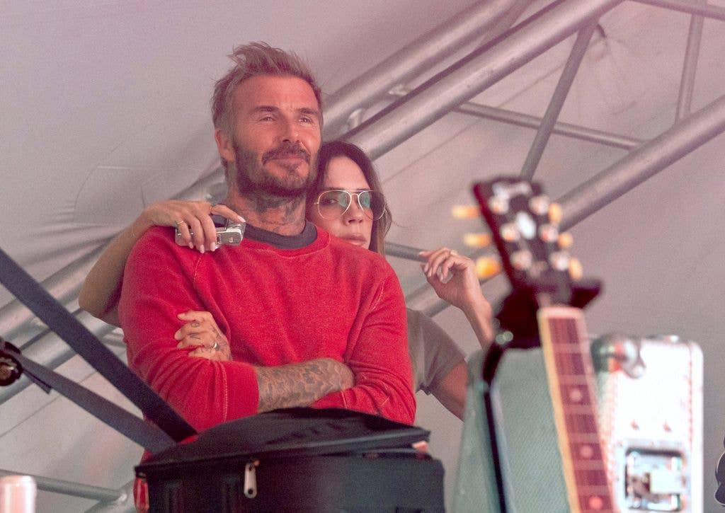 David Beckham in a red sweater stands with arms crossed, holding a camera, next to a woman in sunglasses.