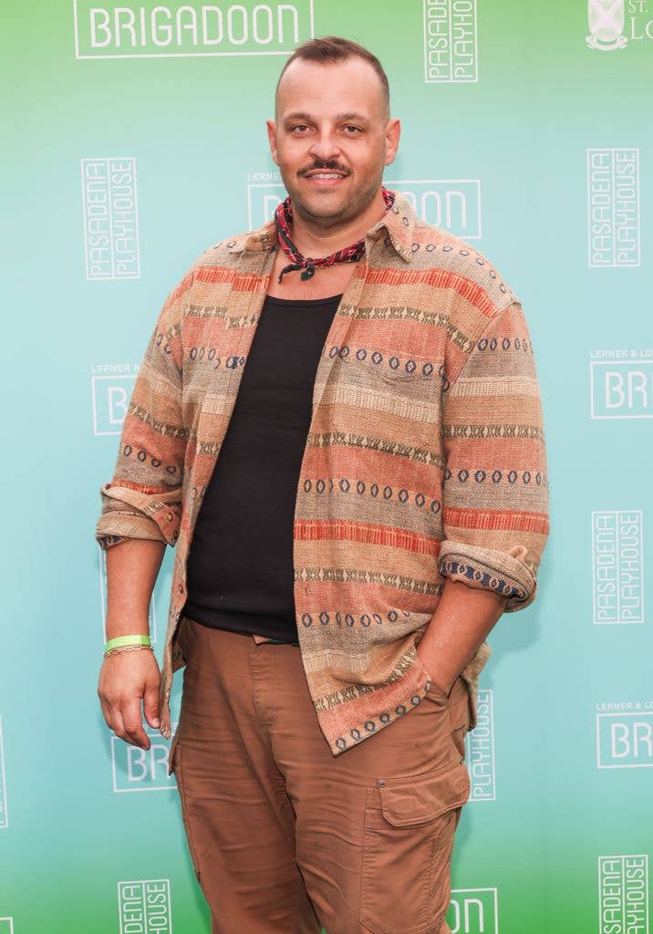 Daniel Franzese stands smiling, wearing a patterned jacket and brown pants, against a green backdrop.