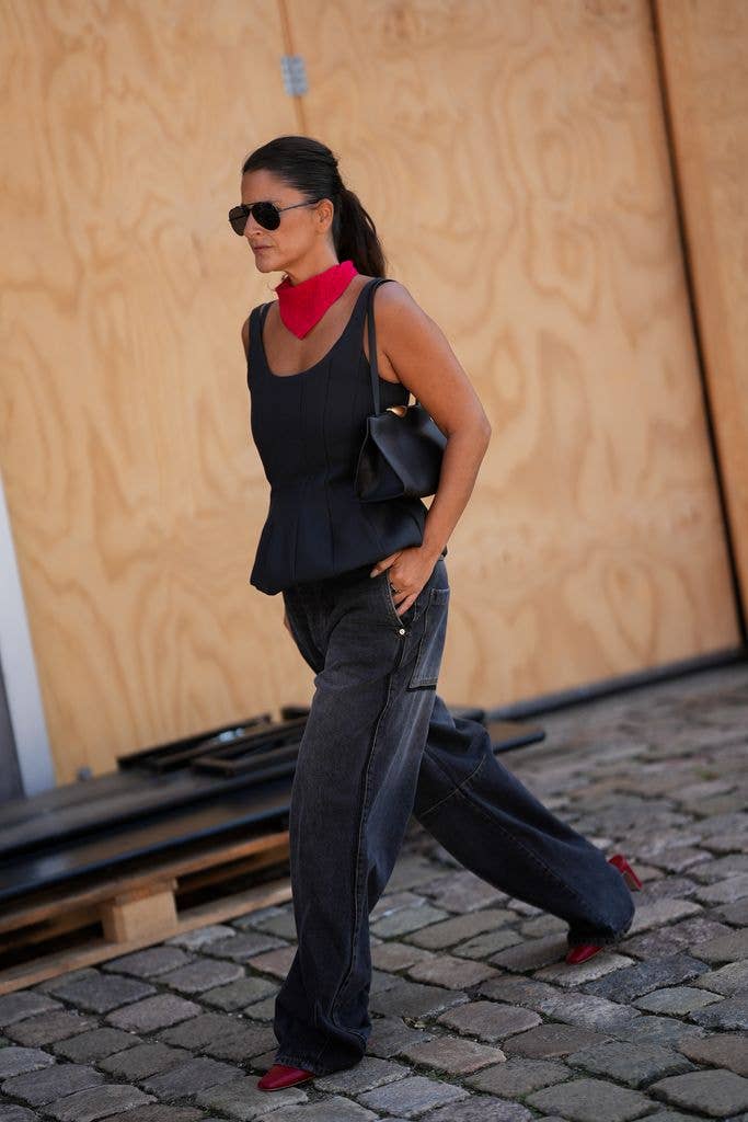  Gili Biegun is seen wearing a black sleeveless peplum top; a bright pink knit neck scarf; dark grey wide leg jeans; red leather pointed toe pumps; a small black leather shoulder bag; aviator sunglasses with dark tinted lenses; dark brown hair worn in a low ponytail during Day Three of Copenhagen Fashion Week