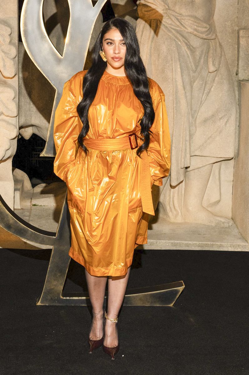 She wore a glossy mustard-yellow belted dress with long sleeves and a high neckline, paired with burgundy pointed-toe heels featuring transparent wedge details.