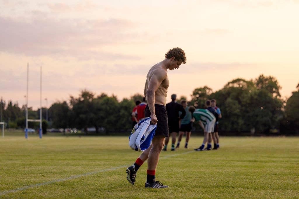 Athlete in black shorts and red socks holds a blue and white bag on a field with other players.