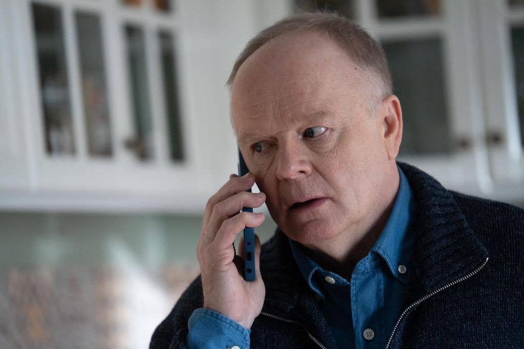 Jason Watkins, wearing a blue shirt and black jacket, talks on a blue phone.