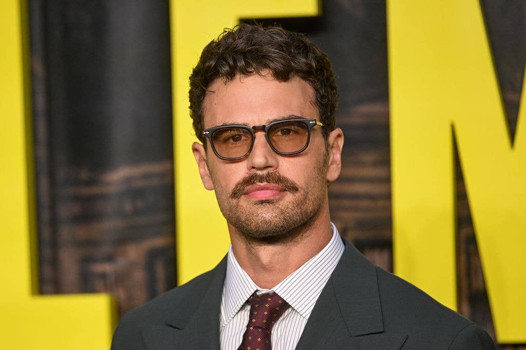 Theo James with a moustache and tinted glasses in front of 'The Gentleman' sign
