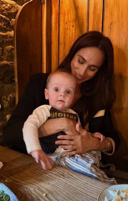 Meghan, Duchess of Sussex, holding a baby in a striped shirt and blue pants.