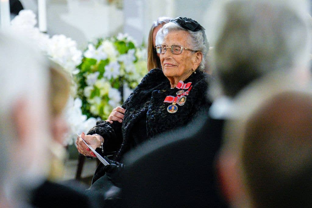 Princess Astrid with glasses and a black fur coat holds a pen, surrounded by others.