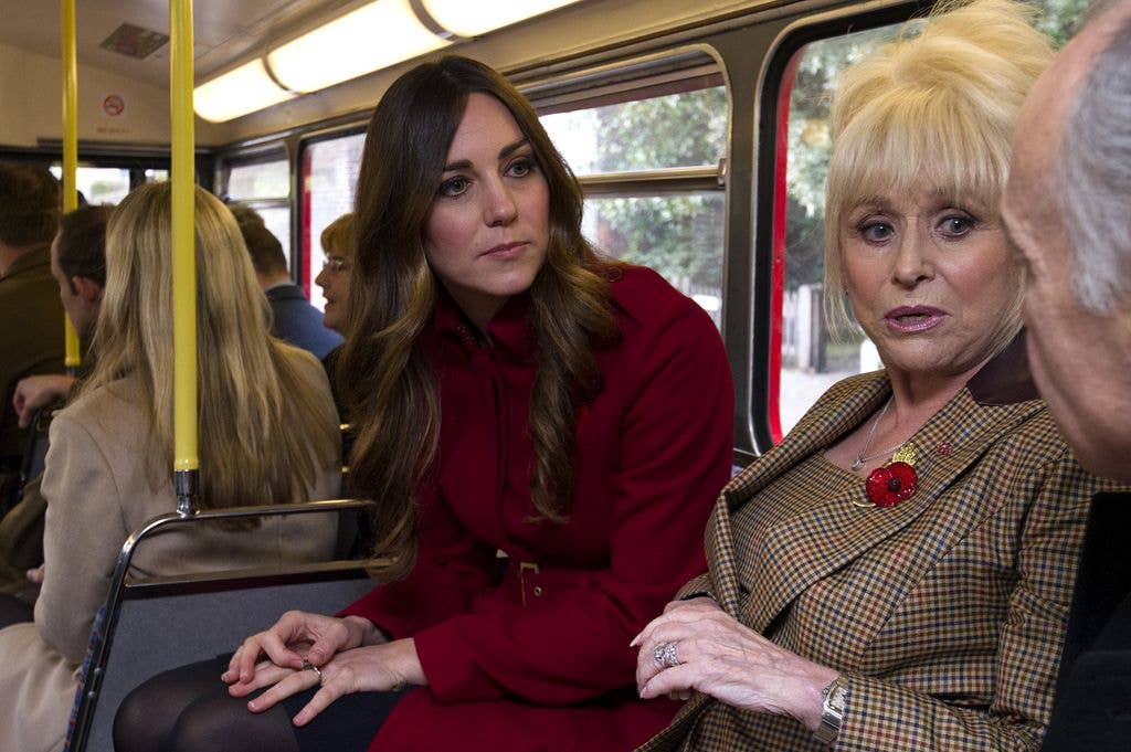 Kate Middleton in red coat and Barbara Windsor in plaid suit sit on a bus.