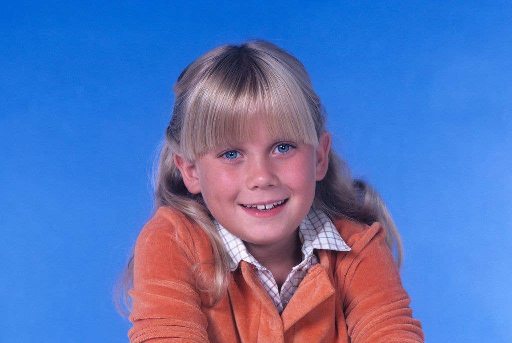 A young girl with blonde hair and bangs, wearing an orange jacket over a checked shirt, smiles for a portrait against a blue background.