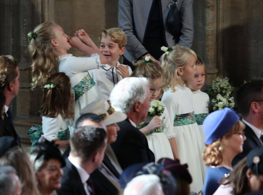 Prince George of Cambridge, smiling and clapping, stands among young flower girls in white dresses.