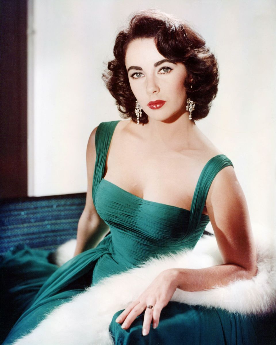 Elizabeth Taylor wearing a green sleeveless low-cut dress, with a white fur wrap on the arm of the armchair, circa 1950