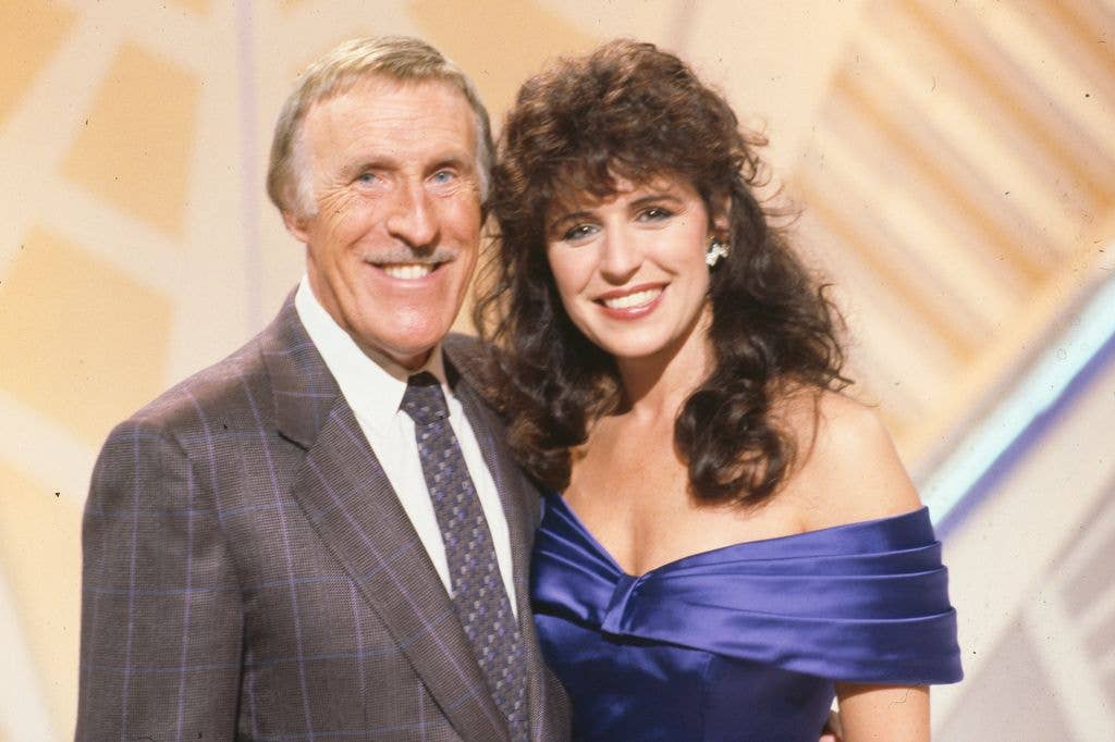 Bruce Forsyth in a checked suit and tie smiles alongside Rosemarie Ford, who wears a blue off-the-shoulder satin dress, both posing for a promotional photo.