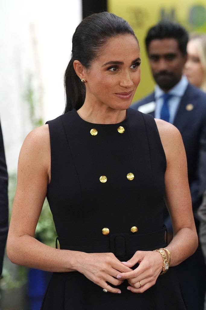 Meghan, Duchess of Sussex, in a black dress with gold buttons, smiling and posing.