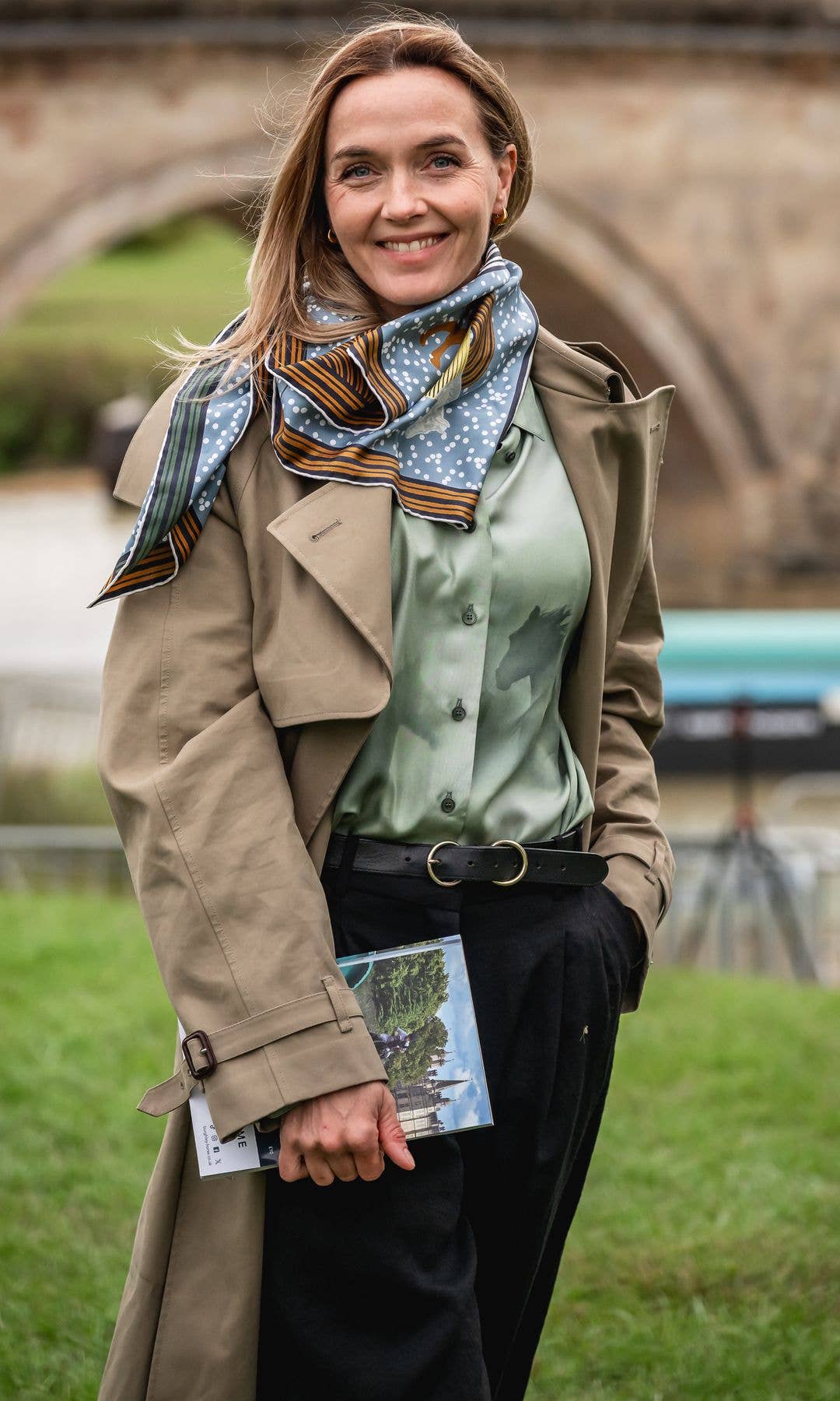 Victoria Pendleton stands in front of a stone bridge, smiling, wearing a trench coat and scarf.