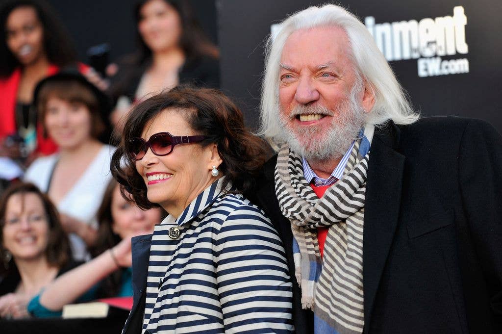 Donald Sutherland, smiling and wearing a striped scarf, stands beside Francine Racette in a striped coat and sunglasses at a red carpet event, with fans visible in the background.