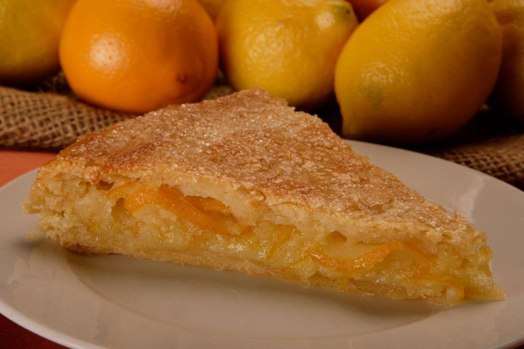 A wedge of lemon tart with a sugar-crusted top and thin slices of lemon visible in the filling sits on a white plate, with whole lemons and oranges in the background.