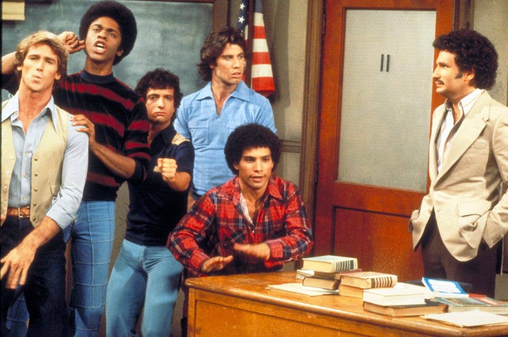 A group of young men in 1970s casual wear stand together in a classroom while a teacher in a beige suit looks on, with an American flag and chalkboard behind them.