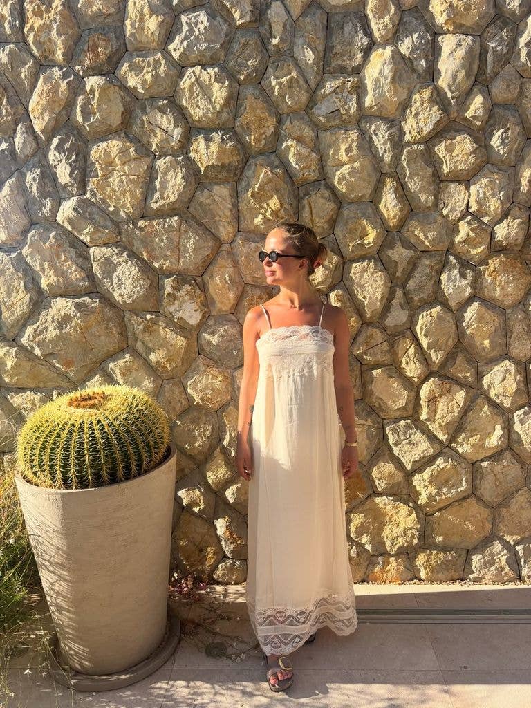 A woman in a white dress stands by a stone wall, wearing sunglasses.