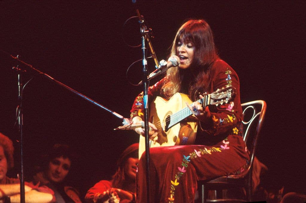 Melanie Safka sings into a microphone while playing an acoustic guitar, wearing an embroidered red outfit, seated on stage in 1974.