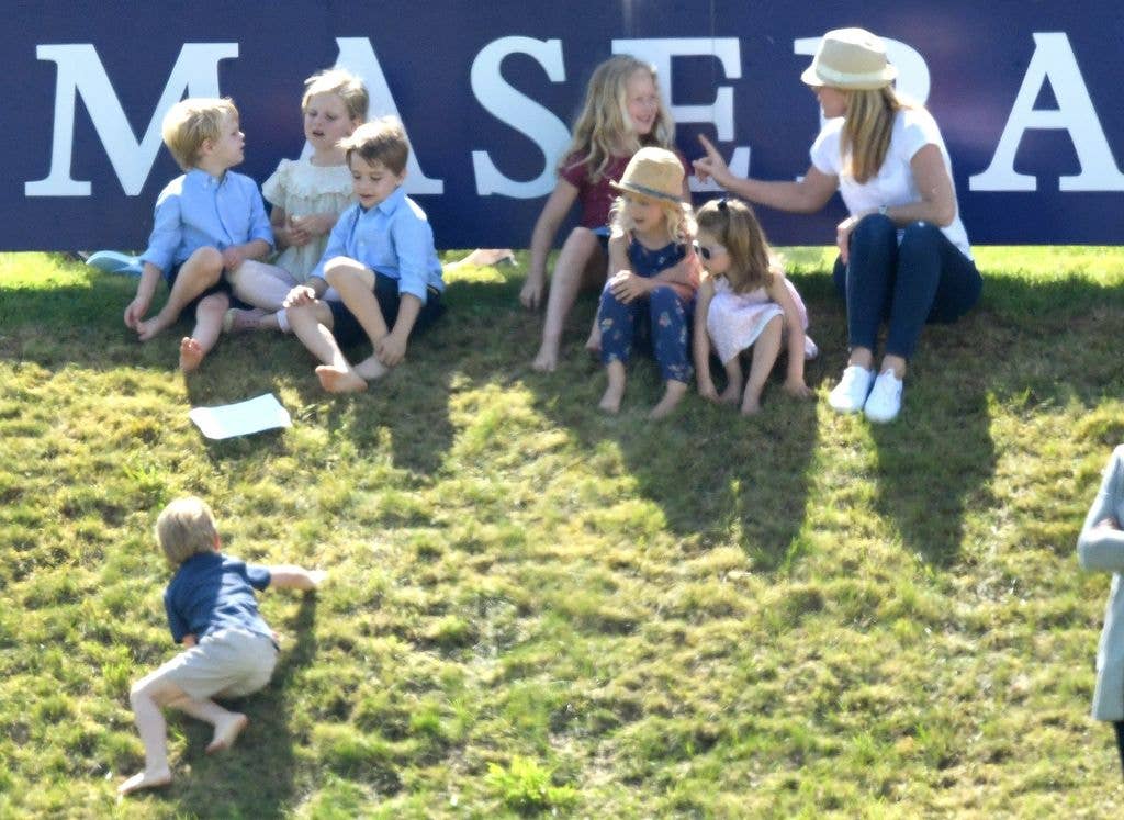 A group of children and a woman sit on grass near a blue banner. One child crawls nearby.