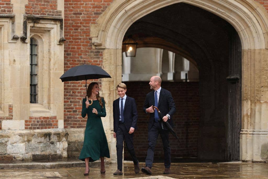 Prince William, Kate Middleton, and Prince George walking under umbrellas in front of a brick building.