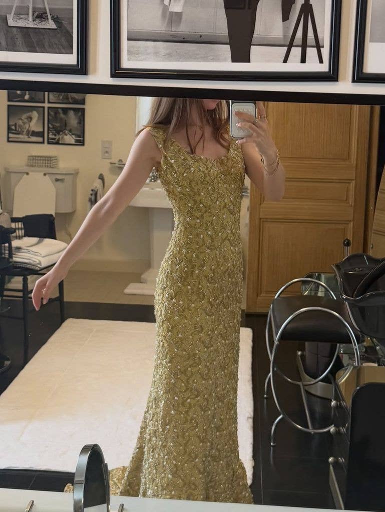 A woman in a gold gown takes a selfie in a bathroom with framed photos on the wall.