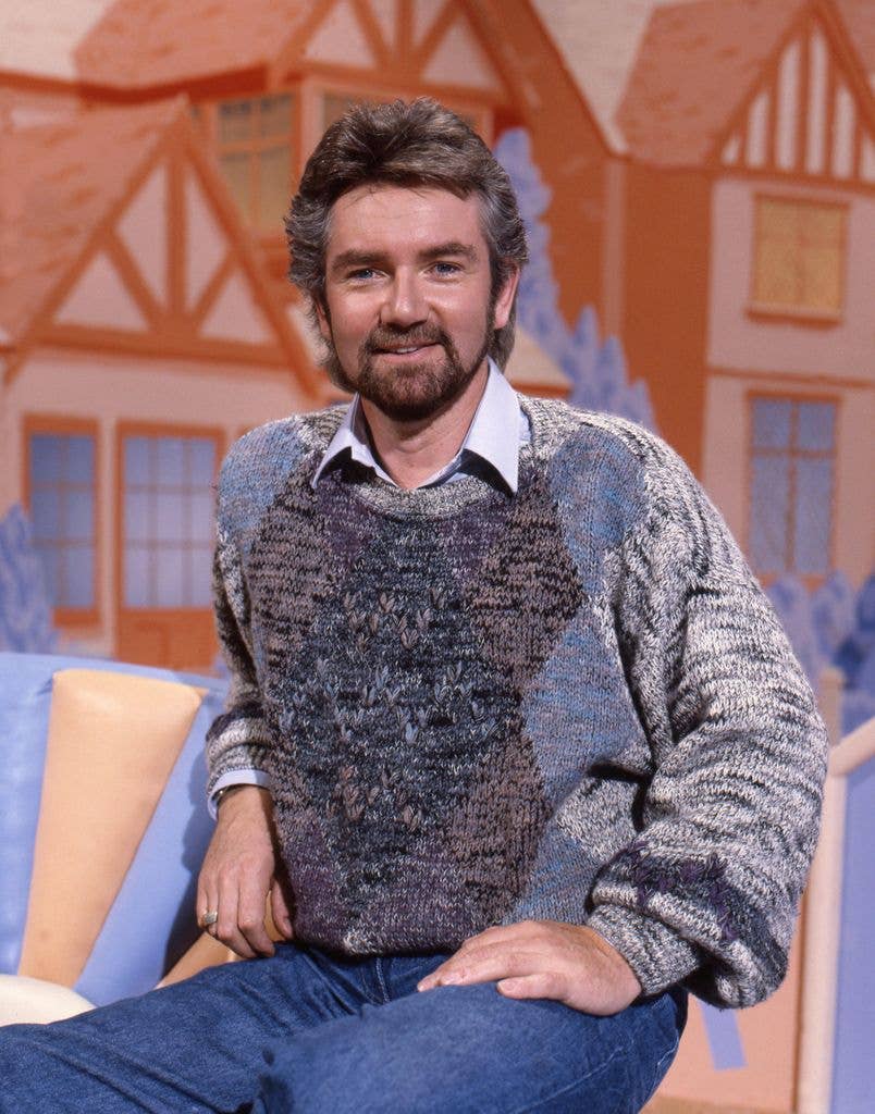 Noel Edmonds, bearded and wearing a patterned knit jumper over a shirt, sits smiling against a painted studio backdrop of Tudor-style houses.
