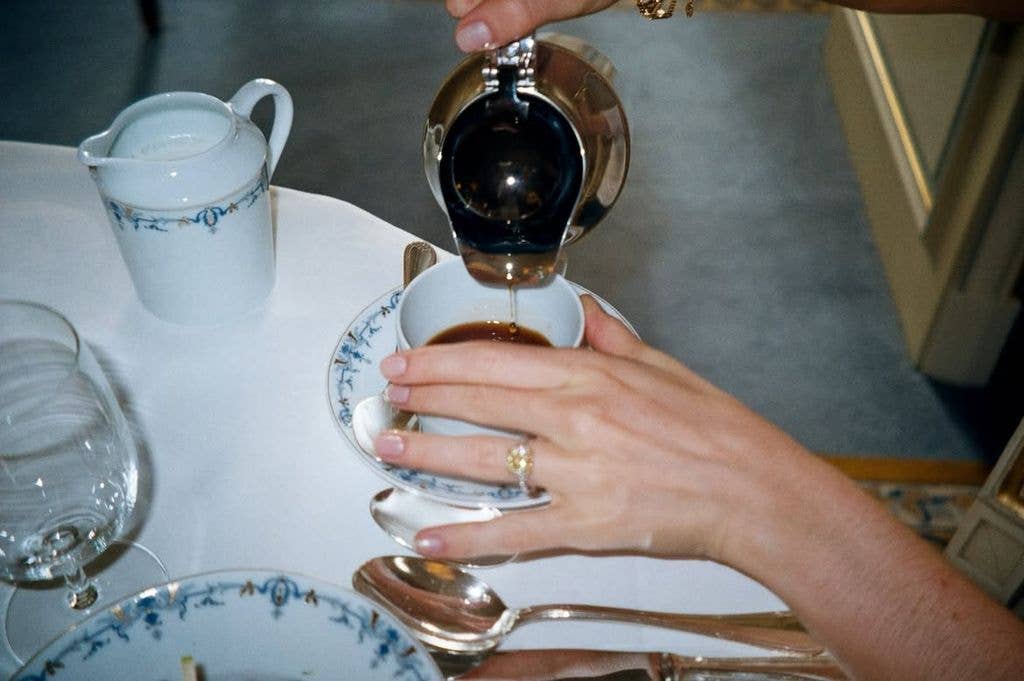 A person's hands pouring coffee into a cup on a saucer, with a ring visible on one finger.