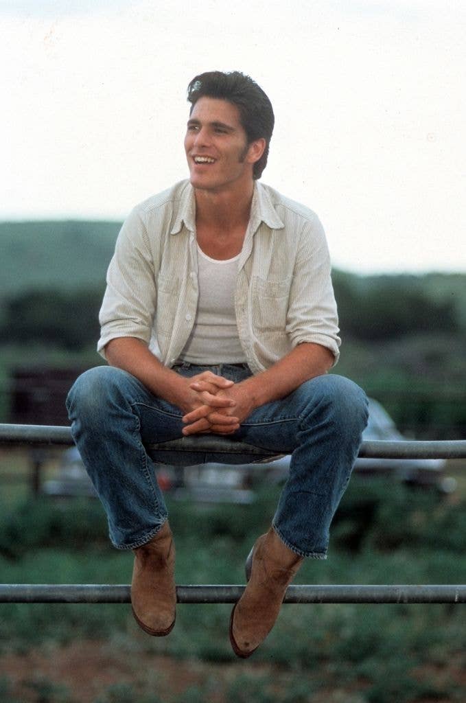 Michael Schoeffling sits on a fence in a white shirt with clasped hands.