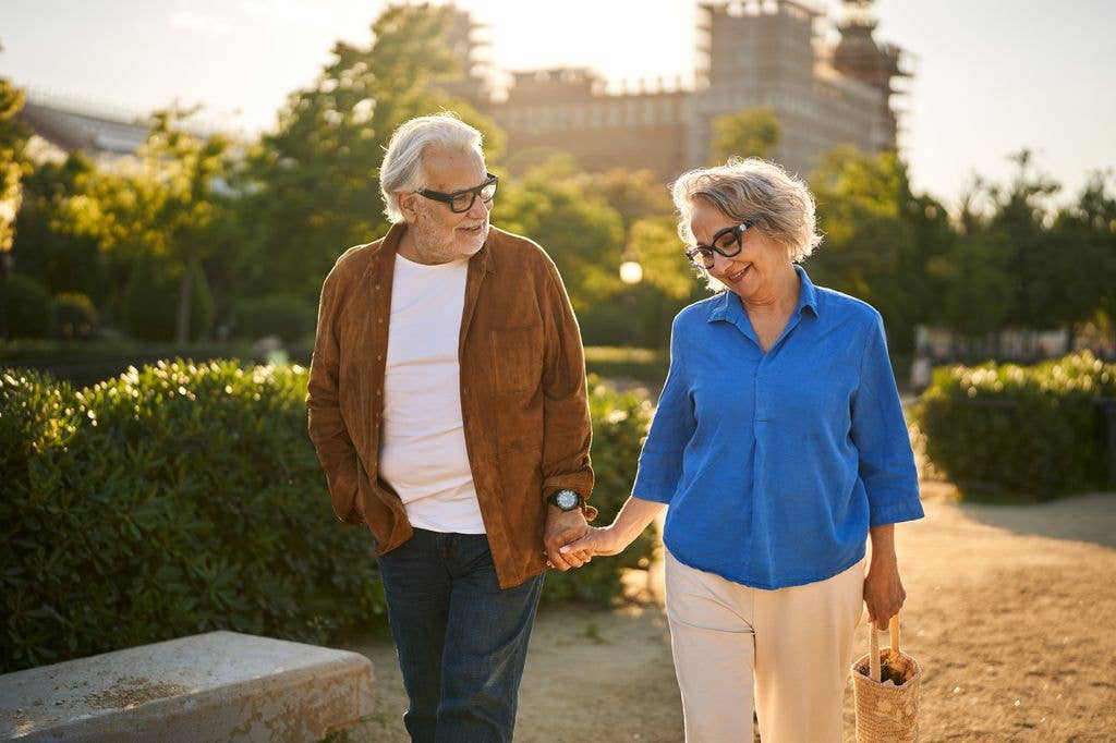 An older man in a brown jacket and an older woman in a blue blouse walk hand in hand along a park path, smiling at each other in golden afternoon light.