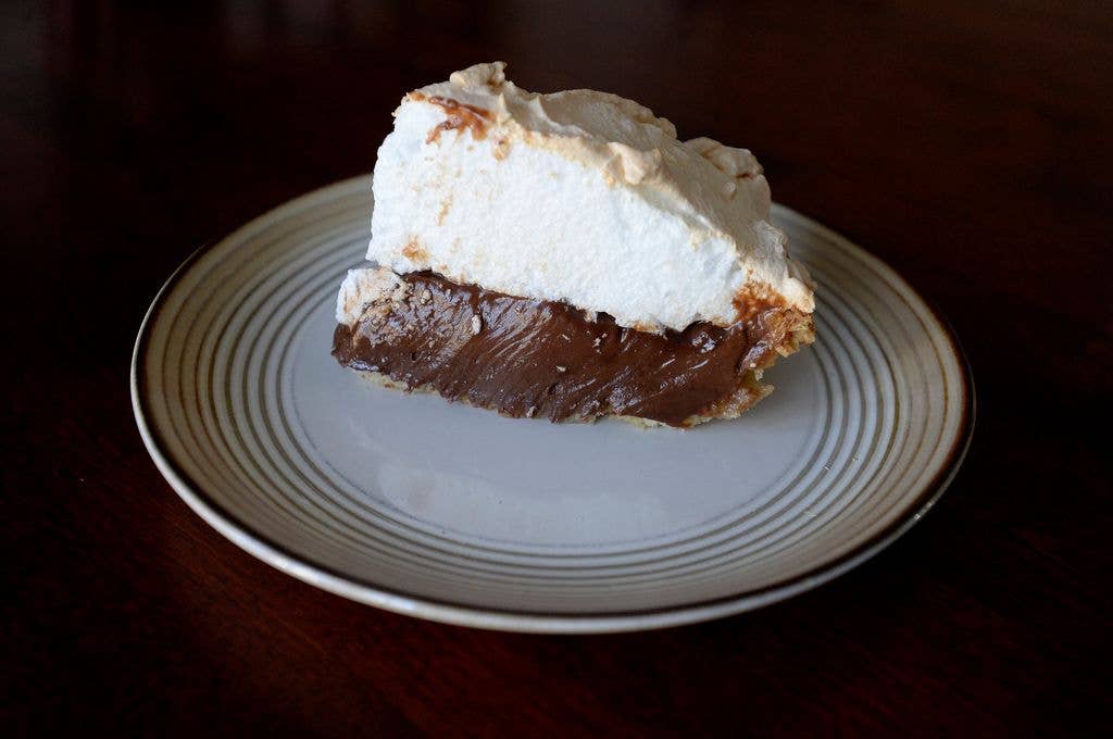 A slice of pie with a rich chocolate custard filling topped with a thick layer of toasted meringue, plated on a striped china dish against a dark background.