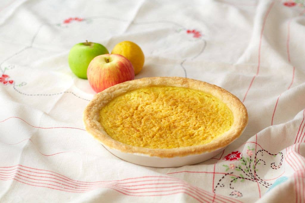 A golden-crusted Marlborough pie in a pale dish rests on a white embroidered tablecloth, with a green apple, a red apple and a lemon arranged just behind it.