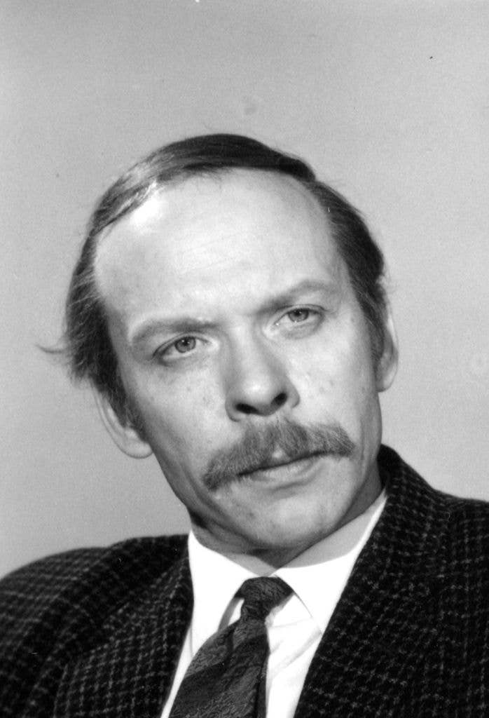 Black and white studio headshot of a man with a moustache, wearing a tweed jacket, shirt and tie, looking directly at the camera.
