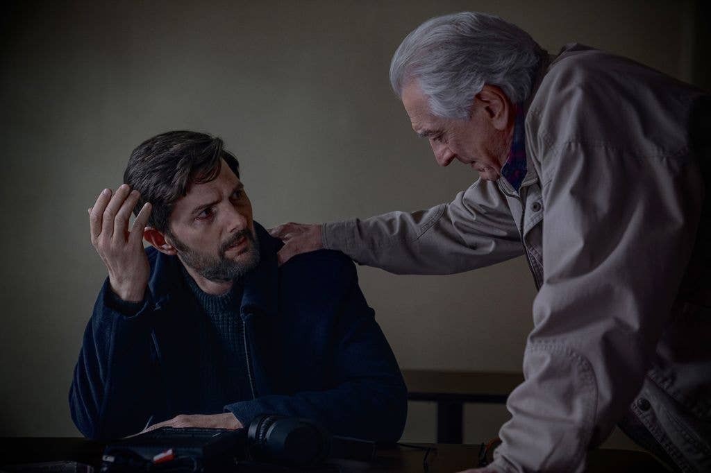 Adam Scott sits at a desk, hand on his head, as an older man places a hand on his shoulder.
