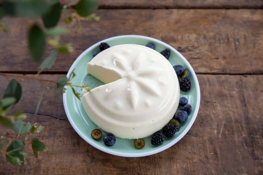 A round, moulded cream dessert with a slice cut out sits on a plate surrounded by blackberries and blueberries, with eucalyptus leaves at the edge of a wooden table.