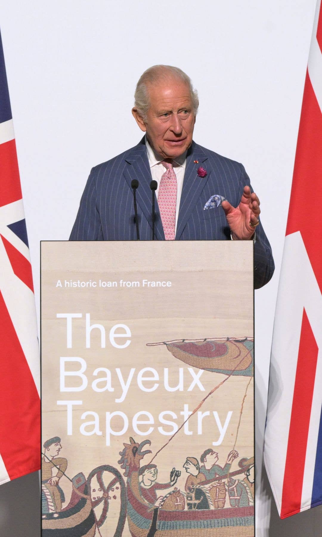 King Charles stands at a podium with flags behind him, speaking.