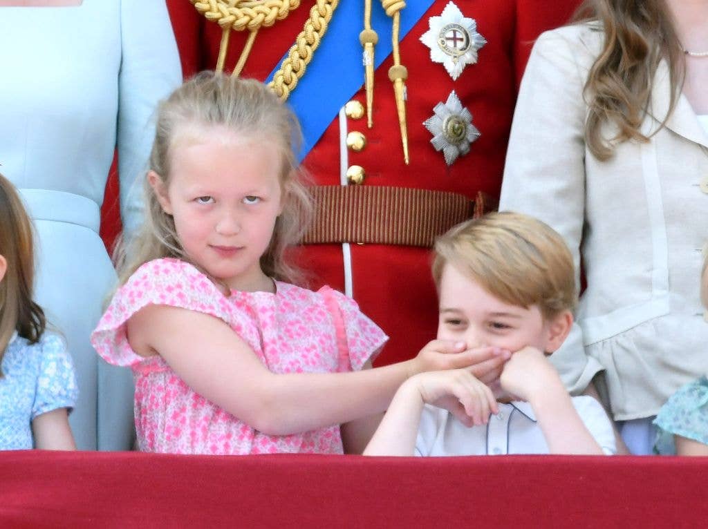 Prince George of Cambridge covers his mouth with his hand while standing next to a young girl.