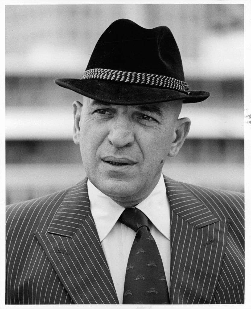 A bald man in a pinstripe suit and fedora looks off to the side with a slight frown, in a black and white photograph.