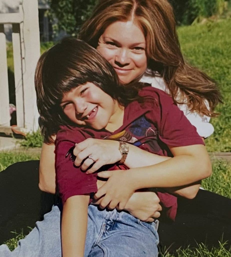 A woman with long brown hair hugs a smiling young boy in a maroon T-shirt while sitting outdoors on grass.