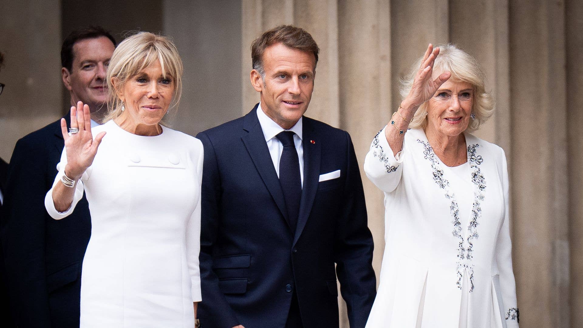 Two women in white dresses and a man in a dark suit, likely at an official event.