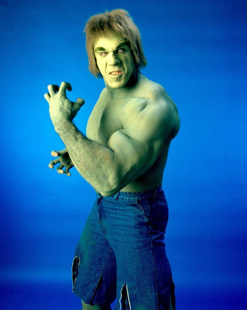 A muscular green-tinted man with shaggy hair snarls and raises a clawed hand, wearing torn denim trousers against a blue studio backdrop.