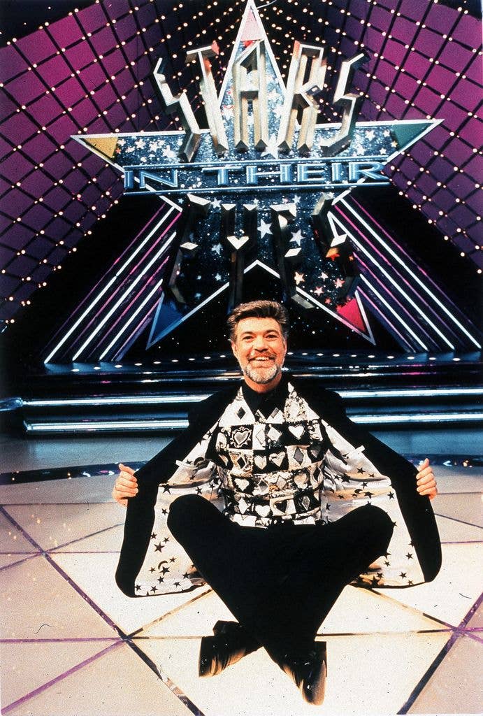 Hans-Peter Reinecke sits cross-legged on the studio floor, smiling and holding open his jacket to reveal a waistcoat printed with playing card suits, beneath the glittering Stars in Their Eyes logo.