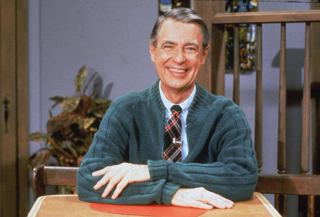 Fred Rogers sits smiling at a table, wearing a green cardigan over a shirt and plaid tie, with a rocking chair and plant in the background.
