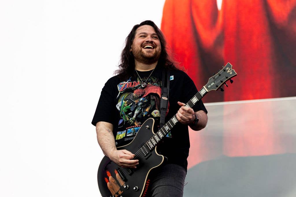 A smiling, long-haired guitarist wearing a black Zelda T-shirt plays a black EVH guitar on stage against a red-lit backdrop.