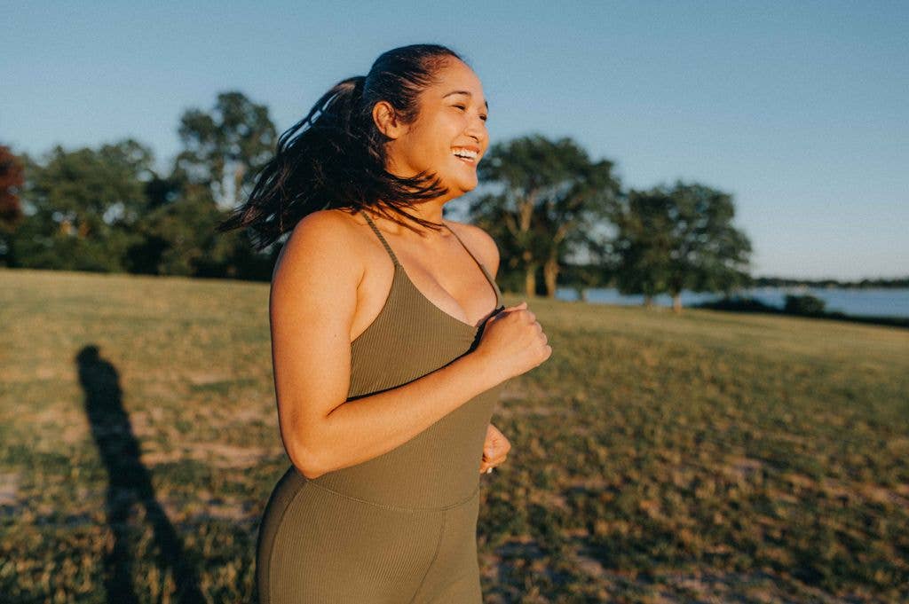 A woman in athletic attire is jogging along a path by a grassy field, with trees and a body of water in the background. The sun casts a warm glow, suggesting it is either early morning or late afternoon.